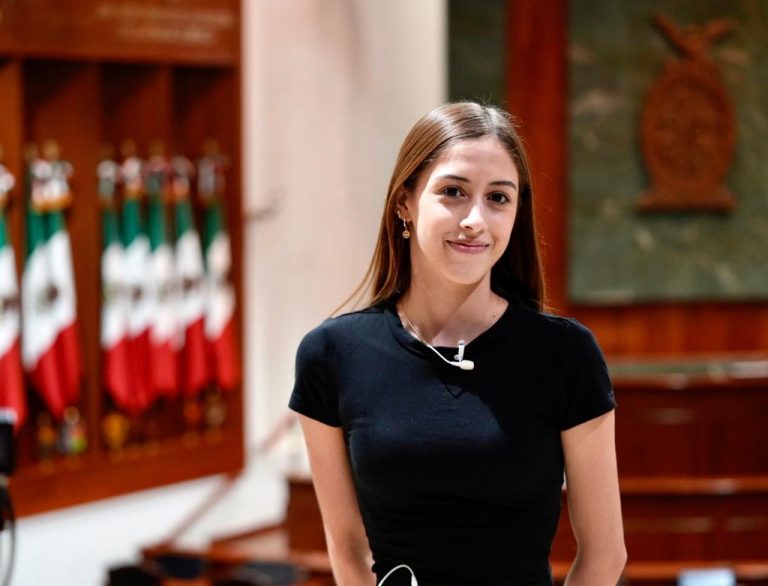 Mariana Chaidez con blusa negra posa para la foto en el Salón de Sesiones teniendo como fondo el gran escudo de Sinaloa tallado en Madera y las banderas de México que adornan el salón.