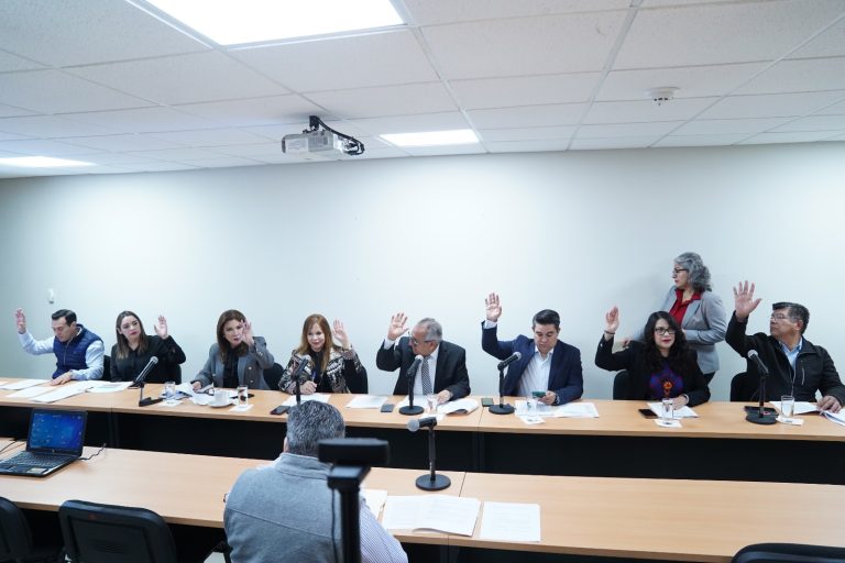 Integrantes de dos comisiones votan levantando su mano en reunión de trabajo en pequeña sala.