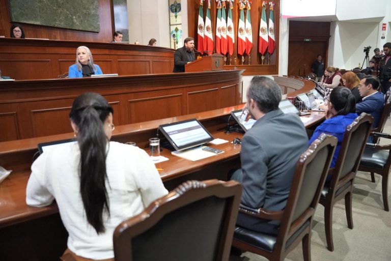Diputada y diputado aparece de espaldas y al fondo la Mesa Directiva y lote de banderas.