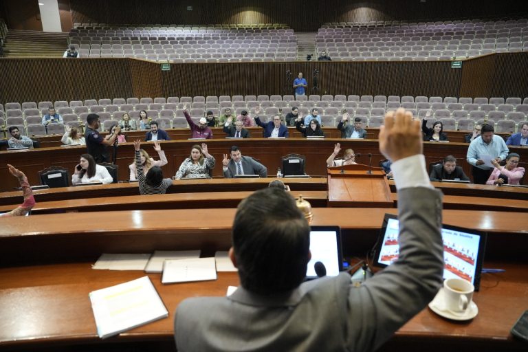 De espaldas aparece presidente de Mesa Directiva. levantando su brazo. Frente a él sus compañeros también votan levandanto su brazo.