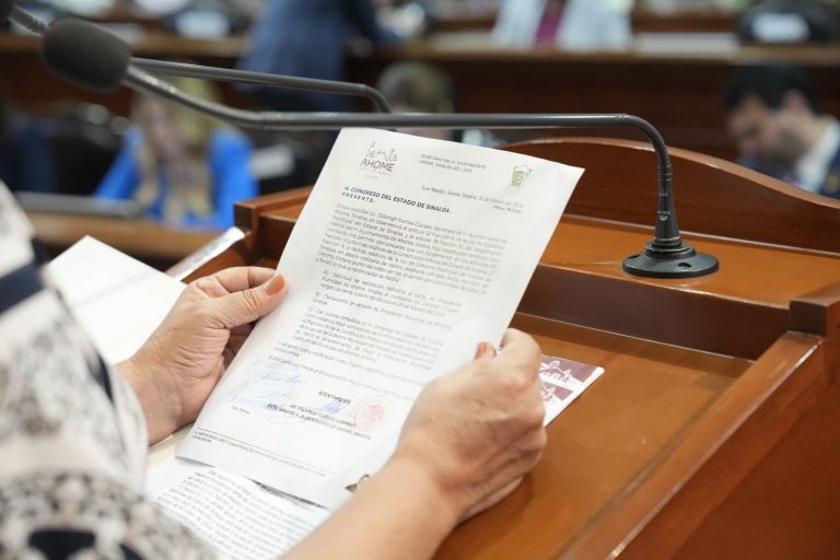 Par de manos sostiene documento que es leído en tribuna.