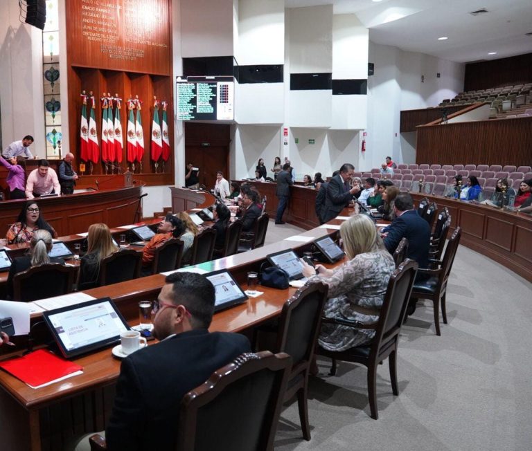 Diputadas y diputados sentados en sus curules y al fondo un lote de banderas de México.