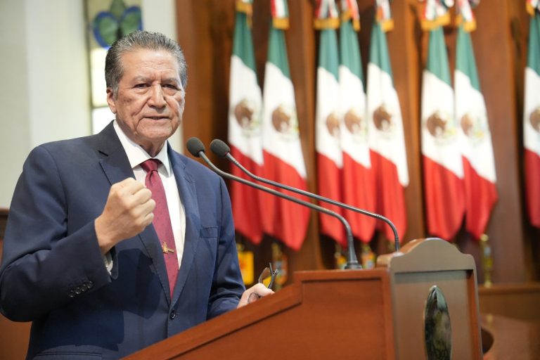 Diputado Feliciano Castro, vestido de traje, habla en tribuna. Tras él un lote de banderas de México.