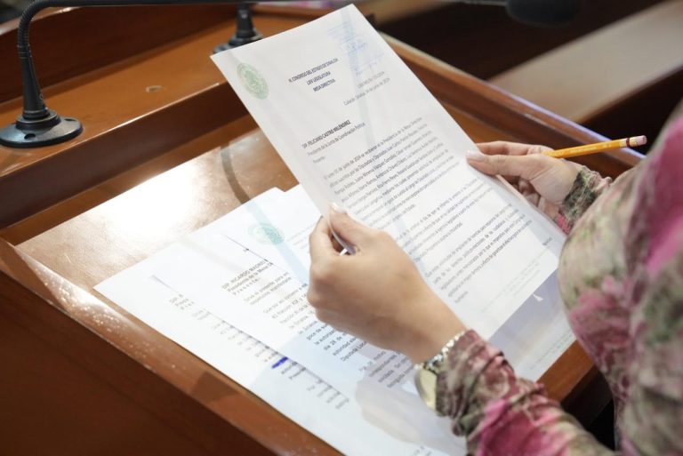 Par de manos de diputada sostiene oficios a los que da lectura ante micrófono en triibuna.