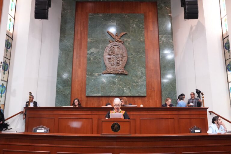 Foto frontal de la Mesa Directiva. Diputada lee en tribuna y al fondo el gran escudo de Sinaloa labrado en madera.