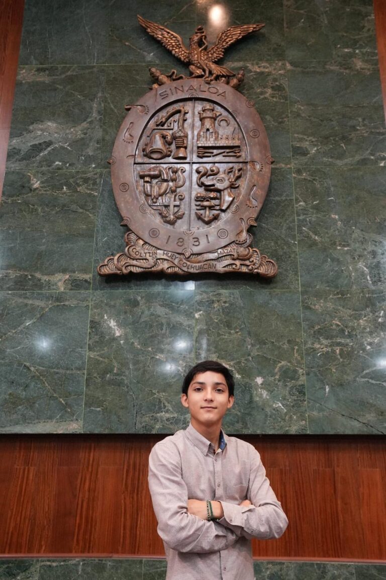 Carlos Emilio Báez posa para la foto en el Salón de Sesiones para el gran escudo de madera, de Sinaloa.