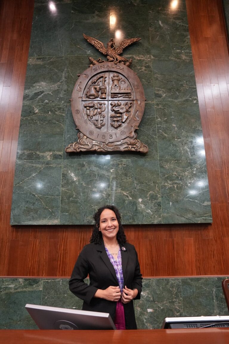 Eunice Andrea Aguirre posa para la foto debajo del gran escudo de Sinaloa, hecho de manera, que está en el Salón de Sesiones.