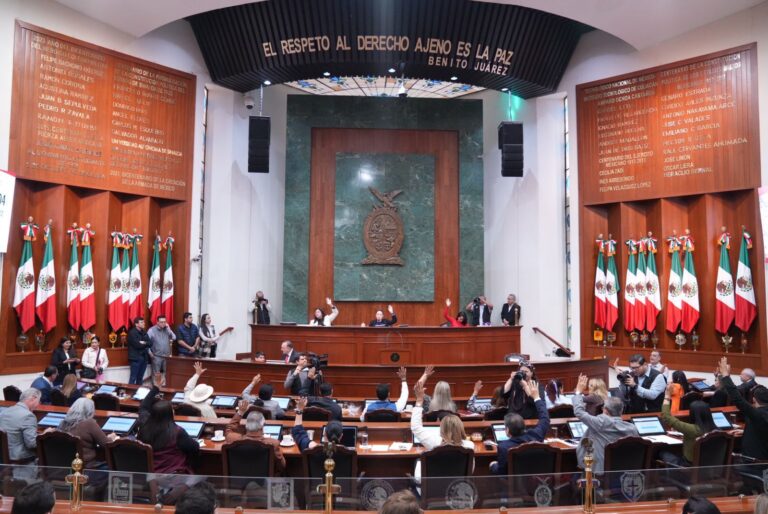 Diputadas y diputados en primer plano, en sus asientos. Al fondo la Mesa Directiva. A los lados lotes de banderas mexicanas.