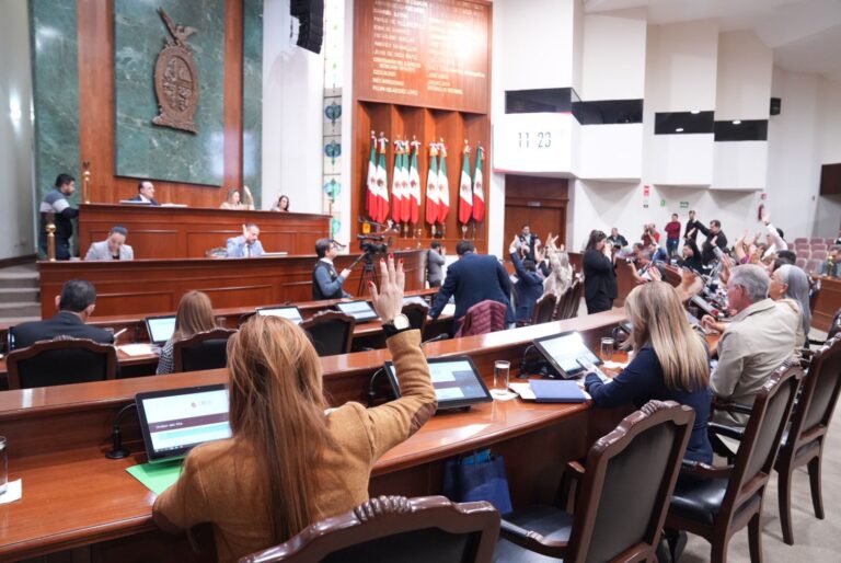 Diputadas y diputados participan en la sesión desde sus lugares. Al fondo la Mesa Directiva y un lote de banderas de México.