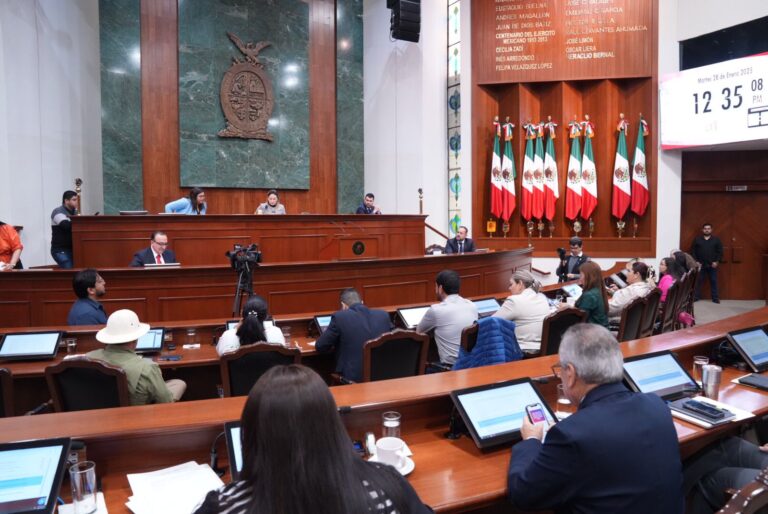 Diputadas y diputados aparecen de espaldas, en sus asientos, pero de frente a quienes forman la Mesa Directiva. Al fondo a la derecha un lote de banderas de México.