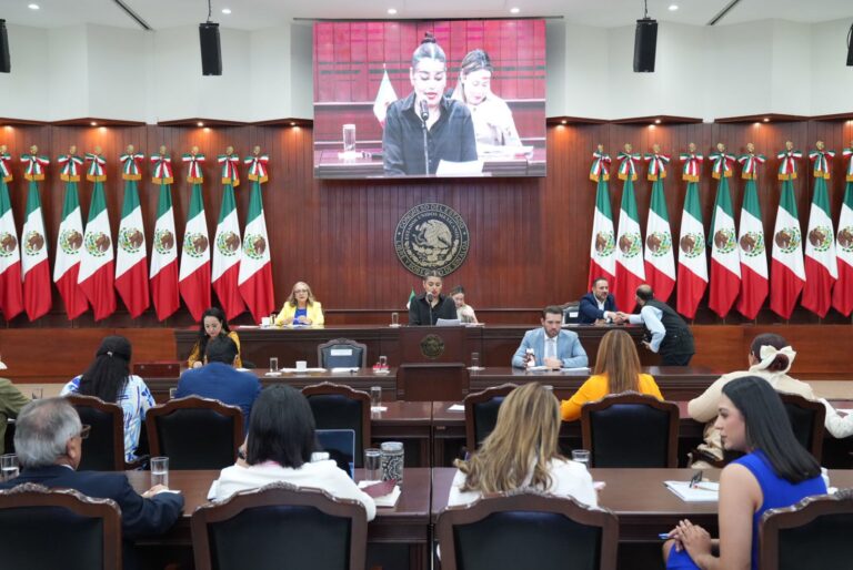 Diputadas y diputados aparecen sentados en primer plano. Enfrente de ellos la Mesa Directiva, pantallas grandes en lo alto y dos lotes de banderas de México.