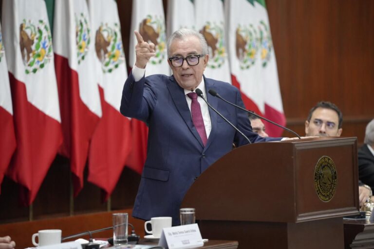 Rubén Rocha habla en podium, vestido de traje y detrás de él hay un lote de banderas de México.