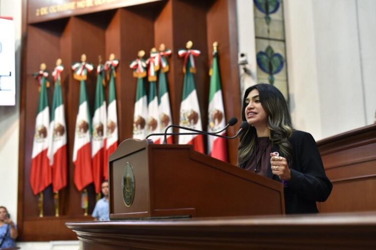 Mujer parlamentaria habla en tribuna. Al fondo un lote de banderas de México.