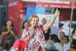 Diputada Tere Guerra levanta la mano izquierda mientras con la derecha sostiene micrófono al hablar en público.