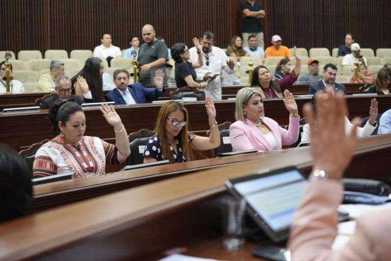 Diputadas en primer plano aparecen sentadas levantando sus manos. Detrás de ellas, compañeras hacen lo mismo.