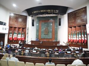 Fotografía amplia del Salón de Sesiones. Diputados al centro. Al fondo la Mesa Directiva. A los costados lotes de banderas de México.